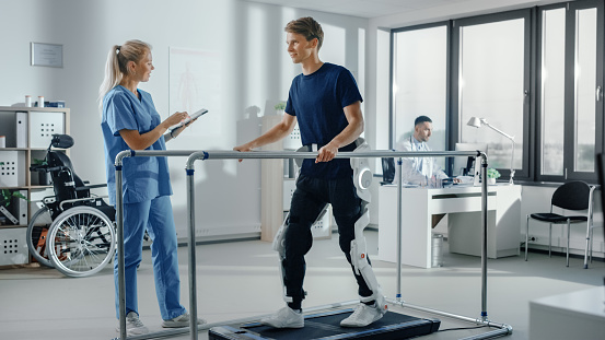 Modern Hospital Physical Therapy: Doctor Uses Tablet Computer, Helps Disabled Patient with Injury Walk on Treadmill Wearing Advanced Robotic Exoskeleton Legs. Physiotherapy Rehabilitation Technology