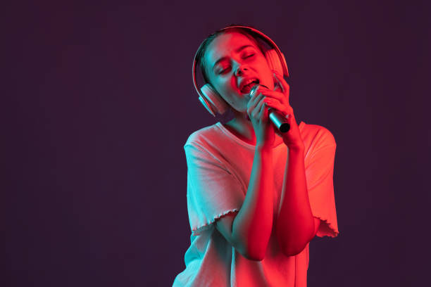 Happy girl with microphone isolated on dark purple studio background in red neon light, filter. Concept of human emotions, facial expression.