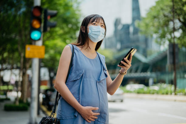 Young pregnant woman with face mask hailing taxi at city street Image of an Asian Chinese pregnant woman with protective face mask ordering a taxi ride with mobile app on smartphone car city urban scene commuter stock pictures, royalty-free photos & images