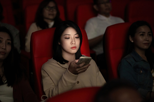 Asian Chinese young woman using smart phone while movie is showing in movie theater. Disturbing other audience around her