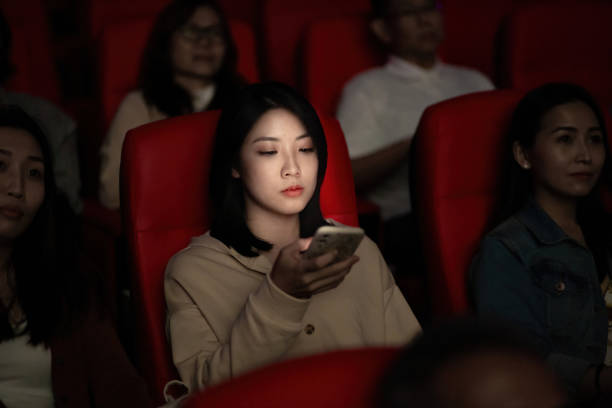 Asian Chinese young woman using smart phone while movie is showing in movie theater. Disturbing other audience around her stock photo