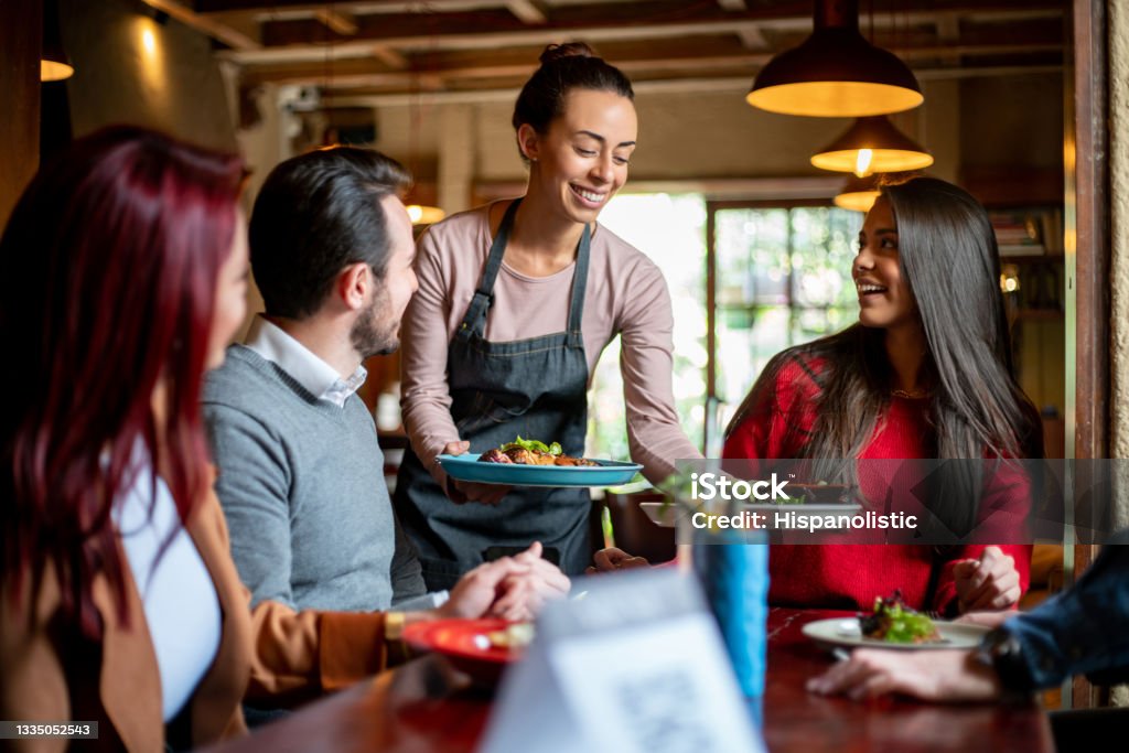 Waitress serving food to a group of customers at a restaurant Happy Latin American waitress serving food to a group of friends having dinner  at a restaurant - food service concepts Restaurant Stock Photo