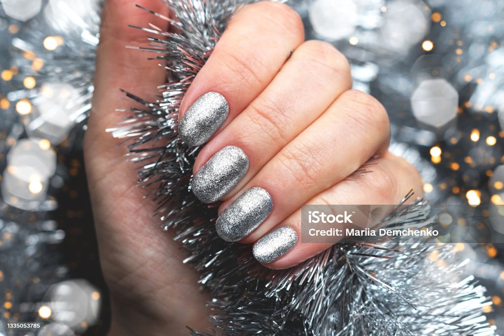 Hand with silver glittered nails on Christmas tinsel background with blurred lights bokeh Female hand with beautiful holiday manicure - silver glittered nails on Christmas tinsel background with blurred lights bokeh Fingernail Stock Photo
