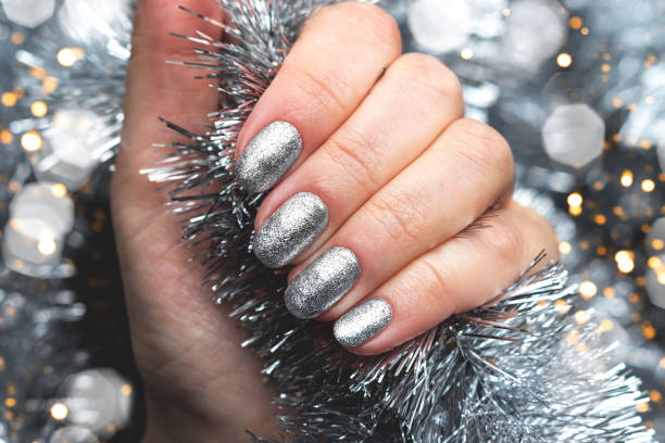 Hand with silver glittered nails on Christmas tinsel background with blurred lights bokeh Female hand with beautiful holiday manicure - silver glittered nails on Christmas tinsel background with blurred lights bokeh christmas nails stock pictures, royalty-free photos & images