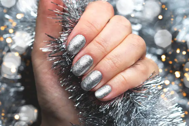 Photo of Hand with silver glittered nails on Christmas tinsel background with blurred lights bokeh