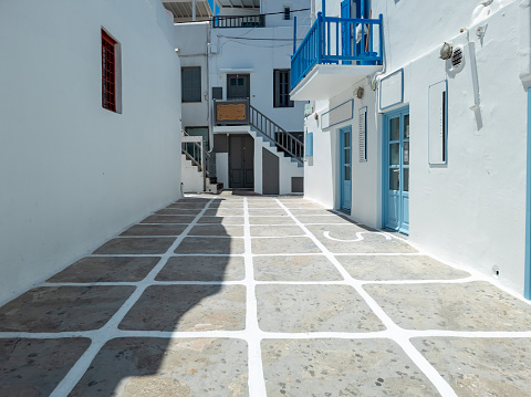 Traditional Greek architecture of whitewashed stonewall buildings closed shops empty cobblestone street. Mykonos island Chora town, Cyclades Greece. Summer vacation Greek resort destination sunny day.