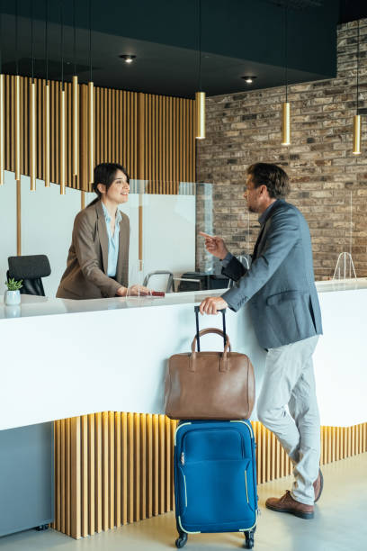 Female Hotel Receptionist Assisting Businessman for Checking In Business man with luggage talking with concierge on hotel reception with sneeze guard protection hotel reception stock pictures, royalty-free photos & images