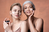 Two female generations mature mother, young adult daughter applying moisturizing sunscreen cream on skin together