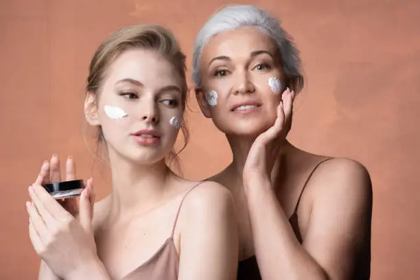 Photo of Two female generations mature mother, young adult daughter applying moisturizing sunscreen cream on skin together