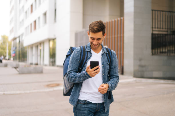 istock Medium shot of cheerful handsome male courier with thermo backpack using navigation app on phone standing in city street 1335065778