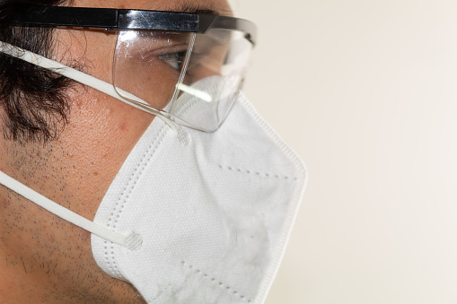Close up of a  hispanic man with long hair wearing a kn95 mask and protective glasses on a white background.