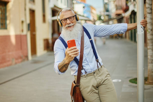 istock Older hipster man using a smartphone and playing music, having fun with mobile phone technology while holding on to a traffic sign in the city - Happy, technological and cheerful senior lifestyle concept - Focus on the face 1335078648