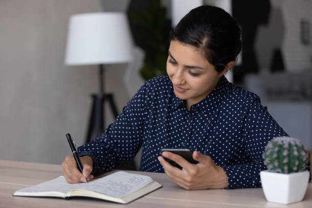 istock Young Indian woman write in notepad use cellphone 1335085299