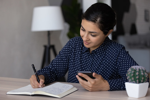Smart Indian woman sit at desk write make note in notebook use smartphone online application. Young biracial female plan handwrite in stationary notepad with cellphone. Time management concept.