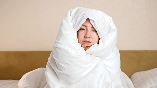 Upset young frozen woman wrapped with soft white duvet sits on large bed in cold room during turning off home heating in winter season