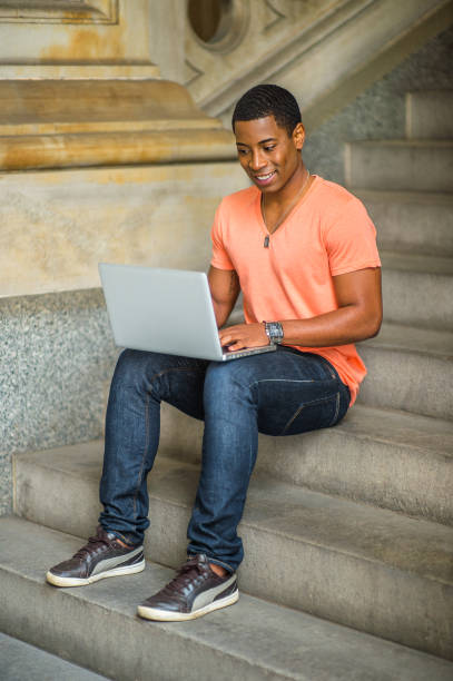 Young man sitting on stairs inside office building, studying on laptop computer