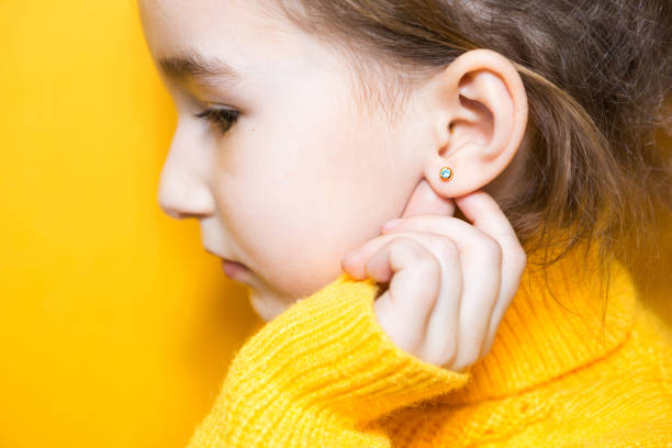 Ear piercing in a child - a girl shows an earring in her ear made of a medical alloy. Yellow background, portrait of a girl in profile. stock photo