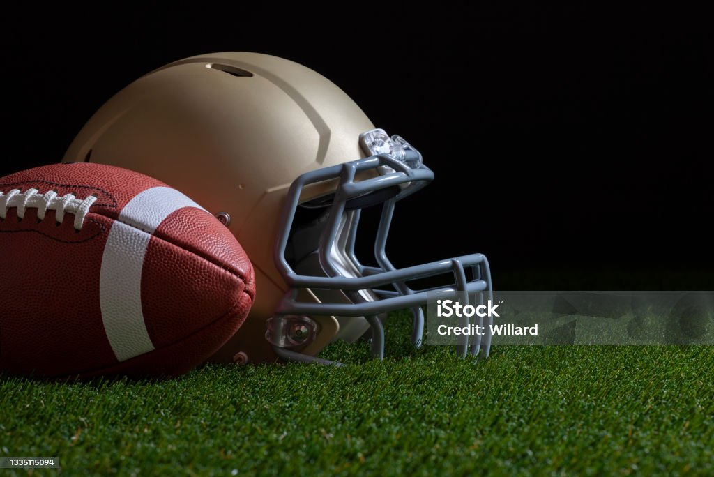 Low angle view of football and gold helmet on grass field with dark background Low angle view of football and gold helmet on grass with dark background College American Football Stock Photo