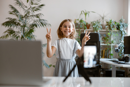 Joyful blonde little girl shows peace gestures posing for mobile phone camera in light room. Recording family blog