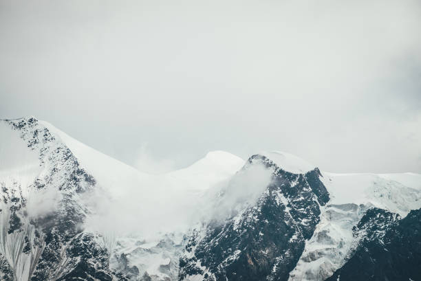 istock Atmospheric mountains landscape with big snowy mountain top with snow cornices in low clouds. Awesome minimal scenery with snow-white high pinnacle in overcast weather. Giant mountain wall in clouds. 1335130637