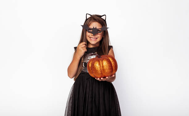 Happy girl with bat mask and shiny pumpkin Cheerful child in black dress wearing cat ears and Halloween paper mask and looking at camera on white background black cat costume stock pictures, royalty-free photos & images