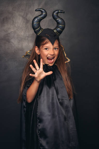Frightening child in black outfit of devil with horns gesturing and shouting on Halloween against black background