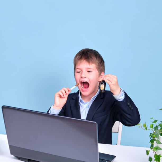 istock A boy in a school suit with a cigarette and alcohol at a table with a laptop, copy space on a blue studio background 1335130945
