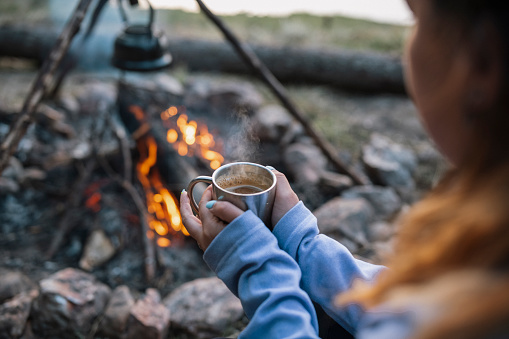 Unrecognisable woman holding hot drink in nature next to the campfire