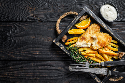 Battered Fish and chips dish with french fries and tartar sauce in a wooden tray. Black wooden background. Top view. Copy space.