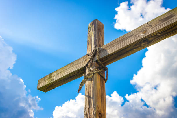 Sunbeam lighting up simple wooden cross with rope sky clouds A ray of sun shines on a wood Christian religious cross, representing Jesus, the Bible and God, with rope from the Crucifixion. Heavenly clouds in the background of a bright blue sky. pentecost religious celebration photos stock pictures, royalty-free photos & images