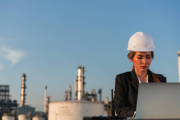 istock Female Engineer In White Hard Hat Wearing Woman's Suit Sitting Looking At Laptop Computer Working Outside,Laptop Lay On Desk In Front Of Petrochemical Industrial 1335167189