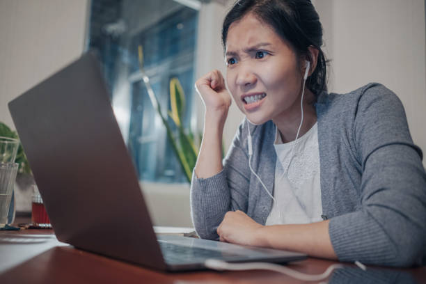 An angry Asian woman having an argument with other people over a video call conference. A medium shot of an angry young adult Asian woman, in a grey cardigan and white t-shirt inside doing video call over a laptop computer on a wood table. She strongly disagrees with the conference. calm before the storm photos stock pictures, royalty-free photos & images