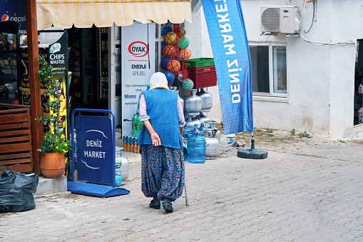 Urla, Izmir, Turkey - June, 2021: Elderly senior woman walking with her cane towards a rural market grocery in Bademler village, Urla, Izmir, Turkey.