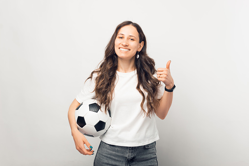 Young gorgeous woman is smiling at the camera, holding a soccer or football ball while showing thumb up over white background.