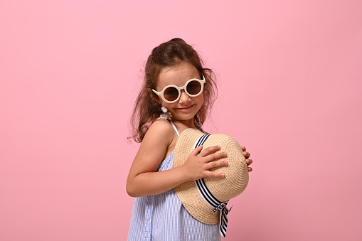 Beautiful shyly baby girl in sunglasses cute smiles posing over pink background with a straw summer hat in her hands. Summer mood concepts with copy space