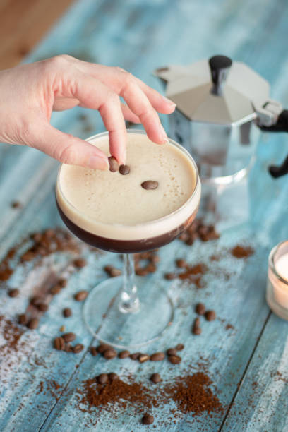 Making Espresso Martini A woman hand garnishing an Espresso Martini in a coupe glass by putting a coffee bean on the top froth layer. The glass is on a blue wooden table and there are coffee beans spread on the table, a mocha pot os seen partially on the back and blurry. coffee liqueur stock pictures, royalty-free photos & images