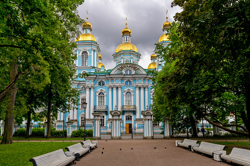 A square with benches in front of St. Nicholas Church in St. Petersburg against a cloudy sky.