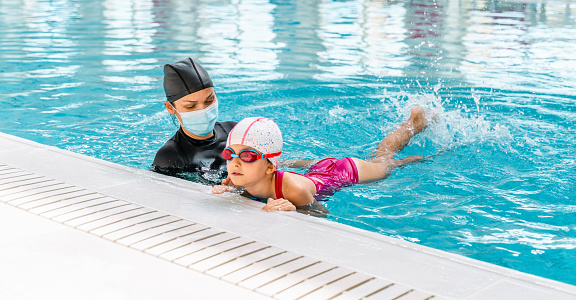Female Coach In Water  with facial medical protective face mask ,Giving child Swimming Lesson In Indoor Pool  in the swimming pool