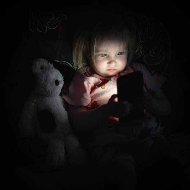 Toddler girl lying on bed and looking on smarthphone screen at night stock photo