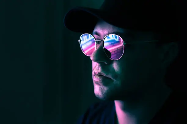 Photo of Neon reflection in glasses. Man in fluorescent light from city led sign. Mysterious cool model in futuristic cyberpunk portrait. Guy in sunglasses. Techno rave party disco art.