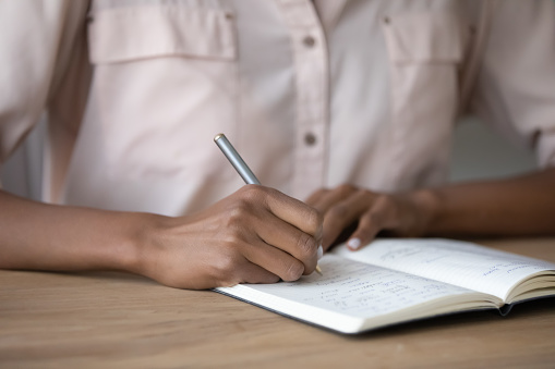 Crop close up of African American woman handwrite take notes in notebook study at home online. Ethnic female write on paper make plans or manage time. Planner, education concept.