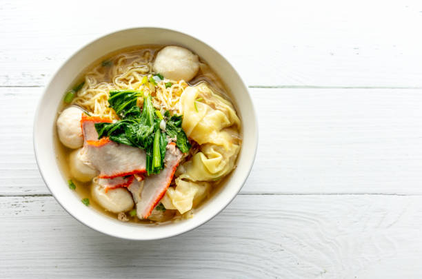 widok z góry wonton i zupy z makaronem w stylu chińskim, polewa kapustą pekińską (bok choy) i czerwoną wieprzowiną na białym drewnianym tle. tajskie jedzenie uliczne - noodle soup zdjęcia i obrazy z banku zdjęć