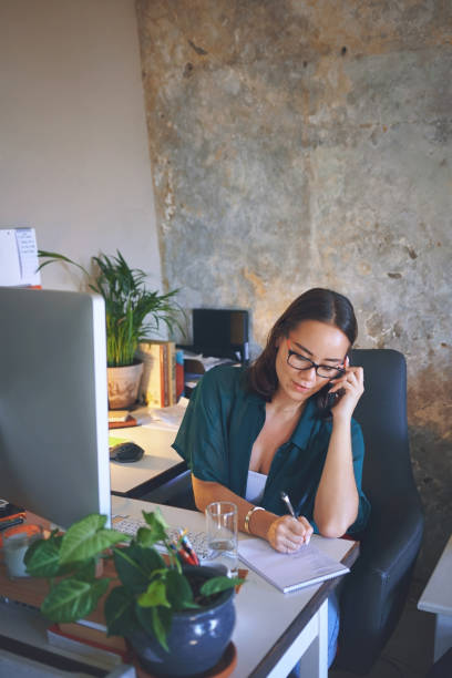 istock Shot of an attractive young woman sitting alone and using her cellphone while writing notes in her home office 1335249716
