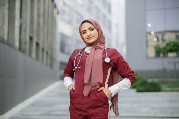 Beautiful Malay lady doctor wearing hijab, scrubs and white coat, outside modern clinic Beautiful Malay lady doctor wearing hijab, scrubs and stethoscope outside modern clinic. Closeup portrait of friendly, smiling confident muslim female doctor in red scrubs, standing outdoors red medical scrubs stock pictures, royalty-free photos & images