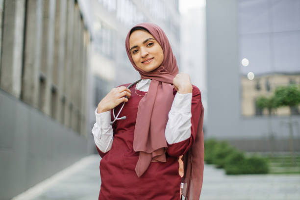 Arab female doctor or medical student, posing and smiling, standing outside modern clinic Arab female doctor or medical student, wearing red medical suit and stethoscope, posing and smiling, standing outside on a background of modern university campus or hospital building red medical scrubs stock pictures, royalty-free photos & images
