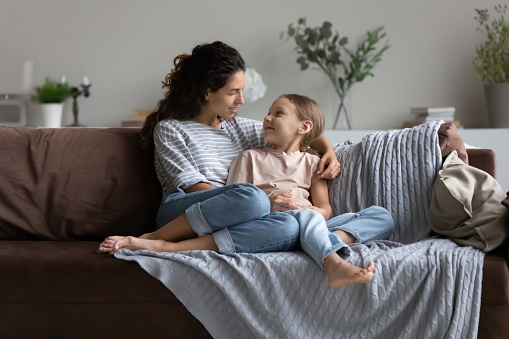 Happy young mom enjoying leisure time with daughter kid at home. Mother sitting on cozy couch, hugging girl, talking to child. Family relationship, motherhood, trust, upbringing concept