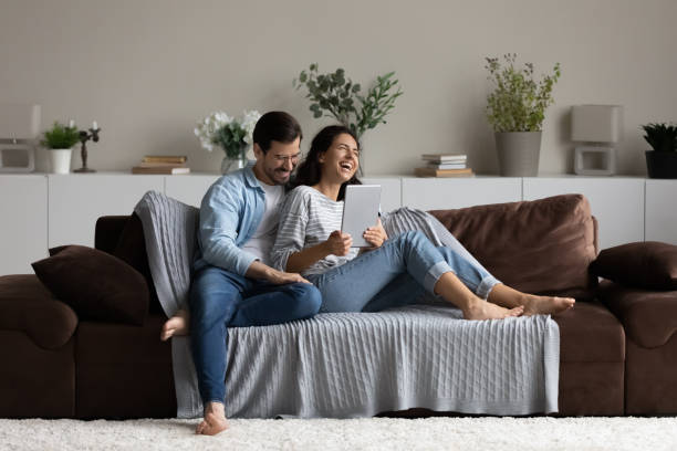 Happy 30s couple relaxing on soft comfortable couch Happy 30s couple relaxing on soft comfortable couch, enjoying leisure time and entertainment in cozy home interior, using tablet, reading book, watching movie, laughing and hugging. Wide shot wide shot stock pictures, royalty-free photos & images