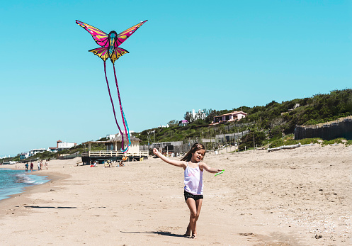 Little girl running with flying kite on the big deserted beach during a travel with family - Little girl playing with kite butterfly on sea shore in a spring sunny day