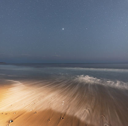 Small waves wash over a beach beneath Summer night skies. Bright Jupiter is seen at center.
