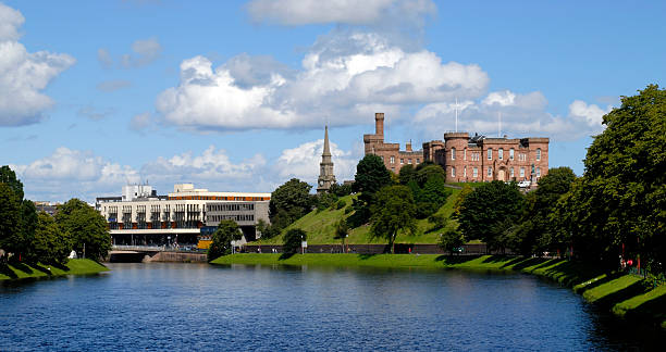 Looking downstream from the Infirmary suspension bridge on a summer day with blue skies and puffy white clouds. In the foreground is the River Ness with its tree lined banks. In the distance is Inverness castle built of local sandstone. Also in the picture are the newly developed apartment overlooking the river and the main road bridge linking the city centre to the west bank of the river